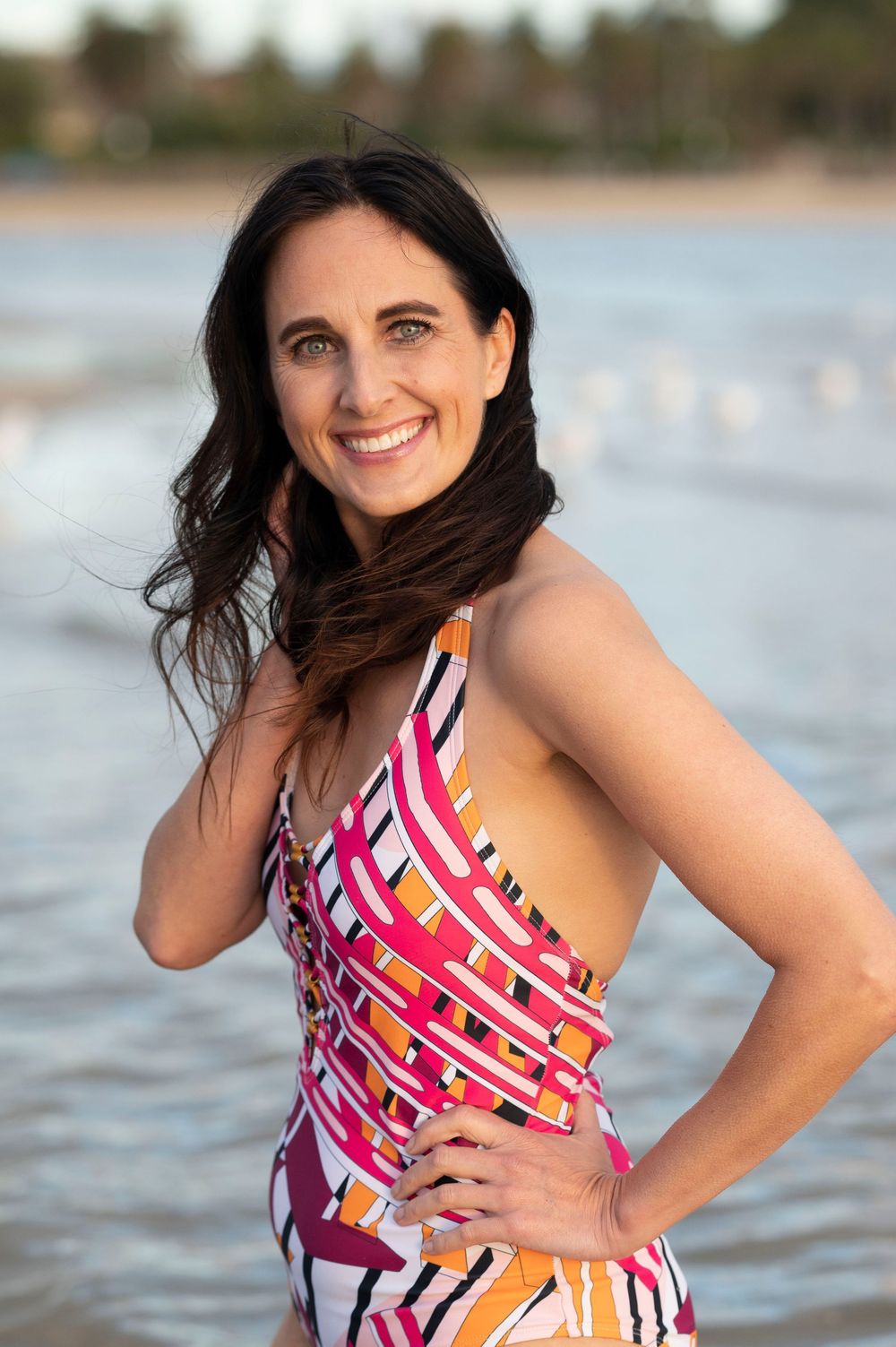 Smiling woman in colorful one-piece swimsuit posing on a sunny beach with ocean behind.