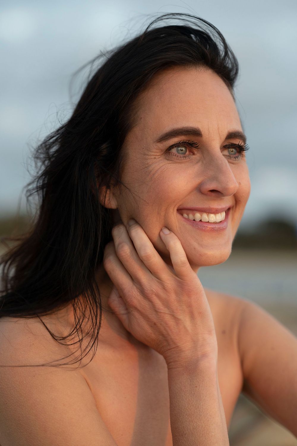Smiling dark-haired woman touching her face at the beach with cloudy sky behind her.