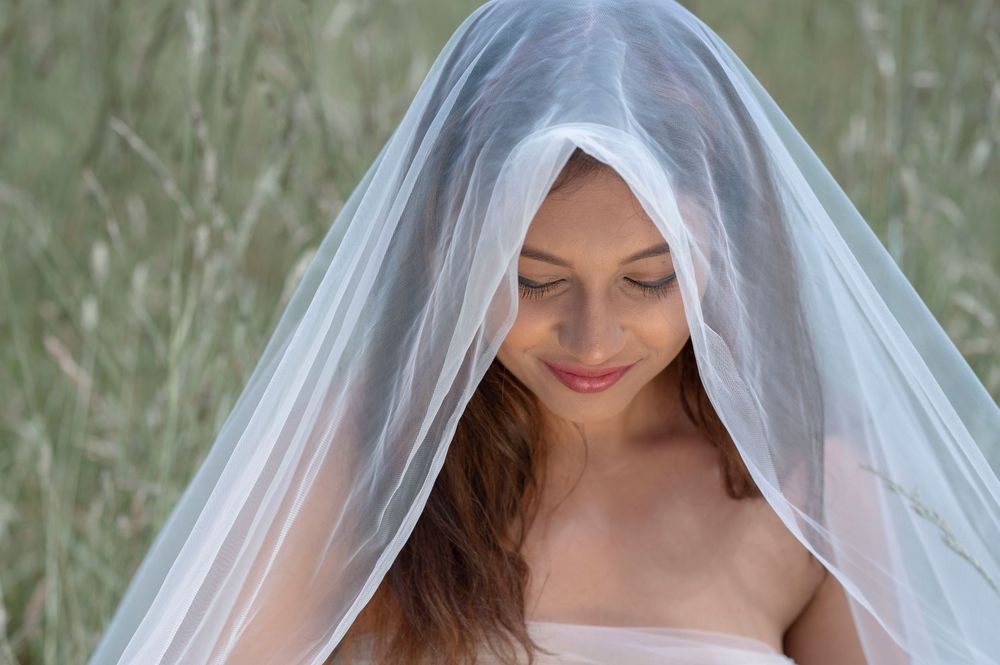 Bride smiling softly under a sheer white wedding veil in a natural outdoor setting.