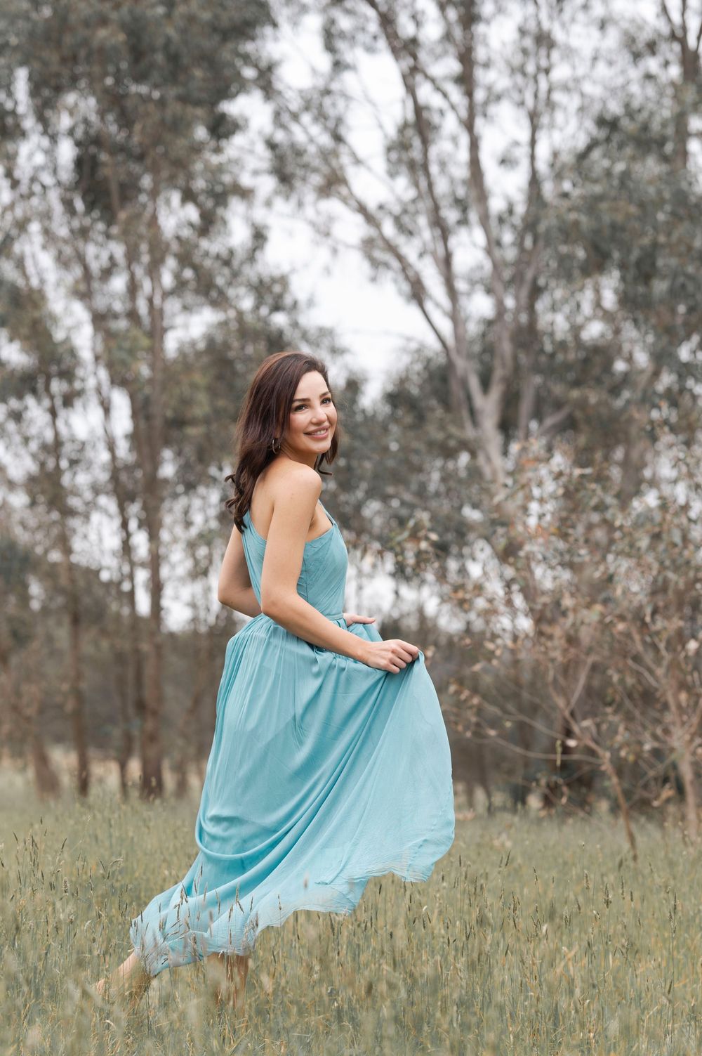 Young woman in a flowing teal dress twirling barefoot in a grassy field with trees.