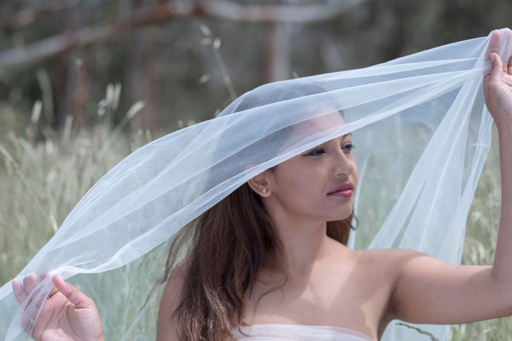 Woman in pink dress holding white veil over her head like a canopy in outdoor field.