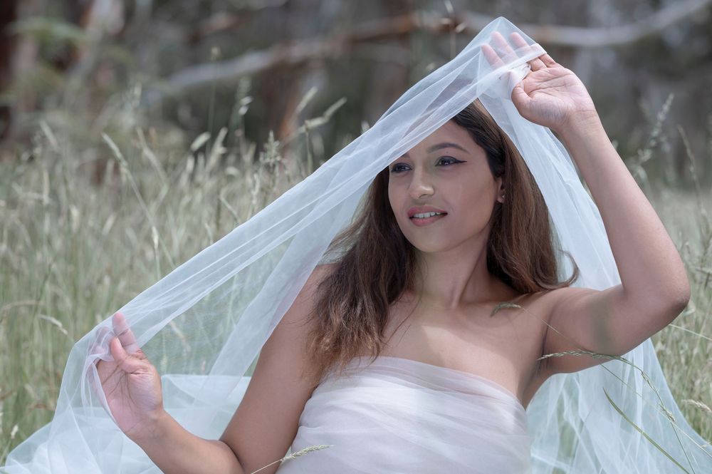 Young woman in white bridal gown lifting veil in outdoor field with tall grass.
