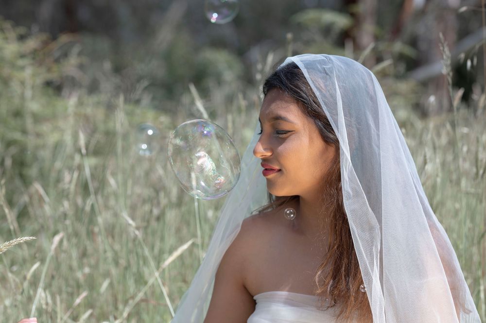 Young bride in white dress and veil smiling as bubbles float around her in a field.