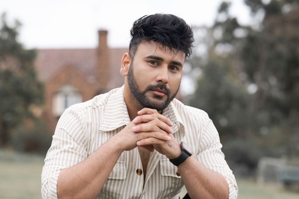 Young bearded man in striped shirt sitting with clasped hands in an outdoor setting.