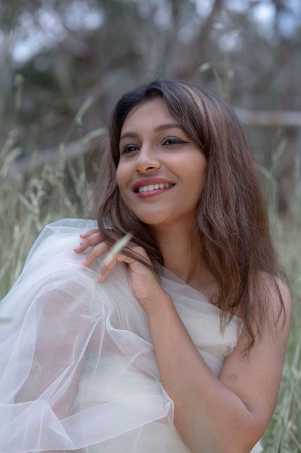 Young woman in white tulle wrap smiling warmly in a soft natural outdoor setting.