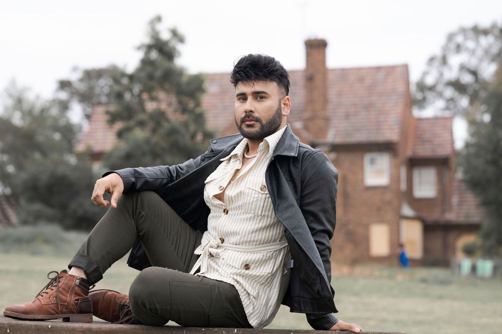 Stylish young man in leather jacket and striped vest sitting outdoors near a brick building.