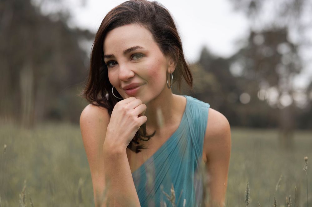A brunette woman in a teal dress smiles thoughtfully in a green field.