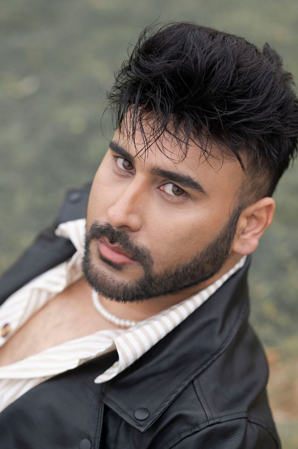 Young man with dark hair and beard wearing a leather jacket and pearl necklace outdoors.