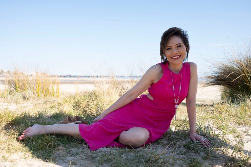 A smiling woman in a pink dress sits barefoot on grass at a sunny beach.