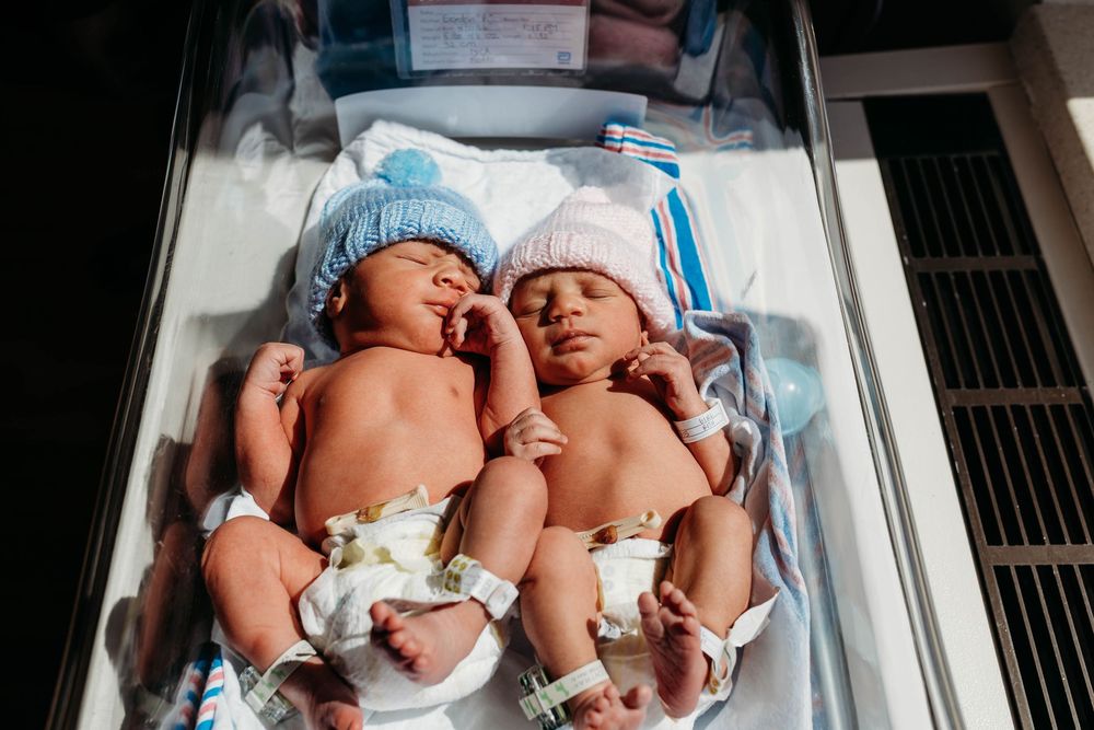 Two newborn twins wearing knit hats resting together in a hospital bassinet under warm light.