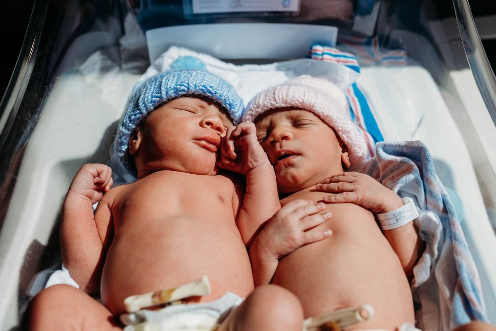 Two newborn twins in a hospital bassinet wearing blue and pink knit hats, sleeping side by side after birth.