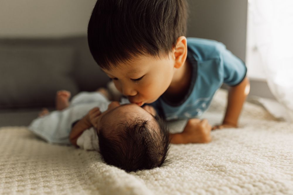 Sweet toddler boy in blue shirt gently kissing newborn baby sibling lying on white blanket on bed.