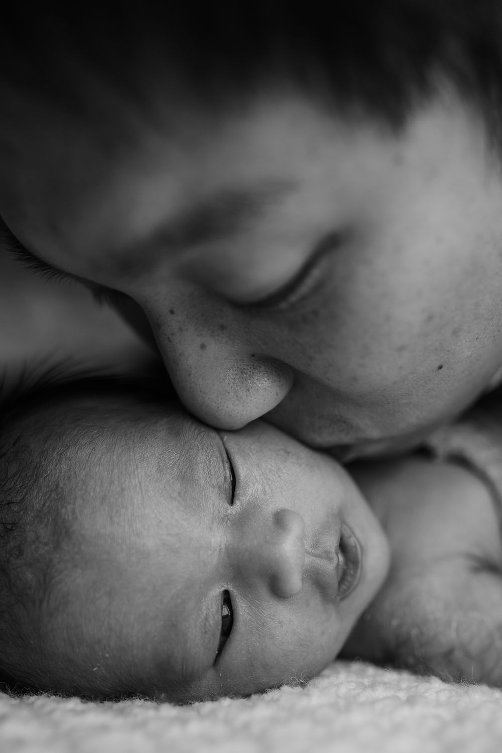 Tender black and white photo of parent lovingly kissing sleeping newborn babys head on soft white surface.