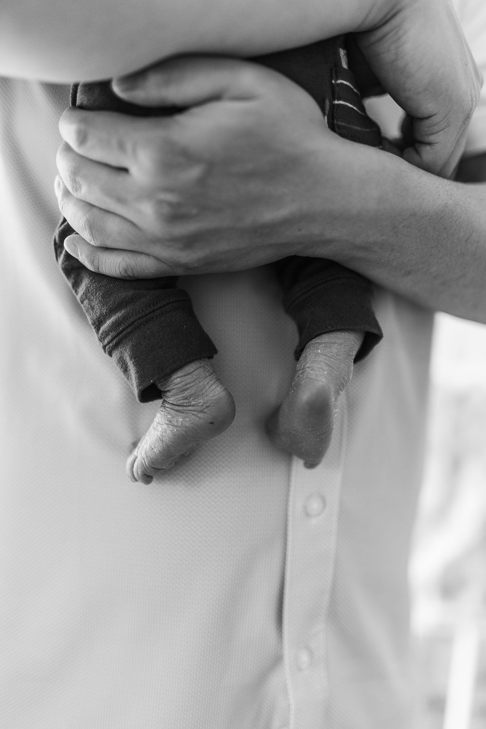 Black and white close-up of parent cradling newborn babys tiny feet and legs against white shirt.