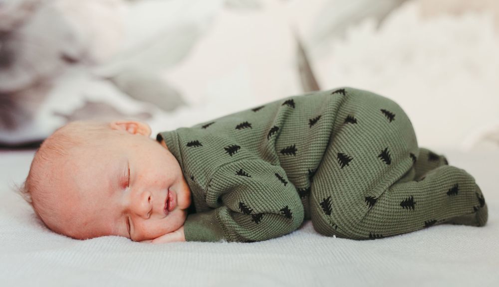 Sleeping newborn baby in a green Christmas tree print footie pajama lying on a soft white blanket.