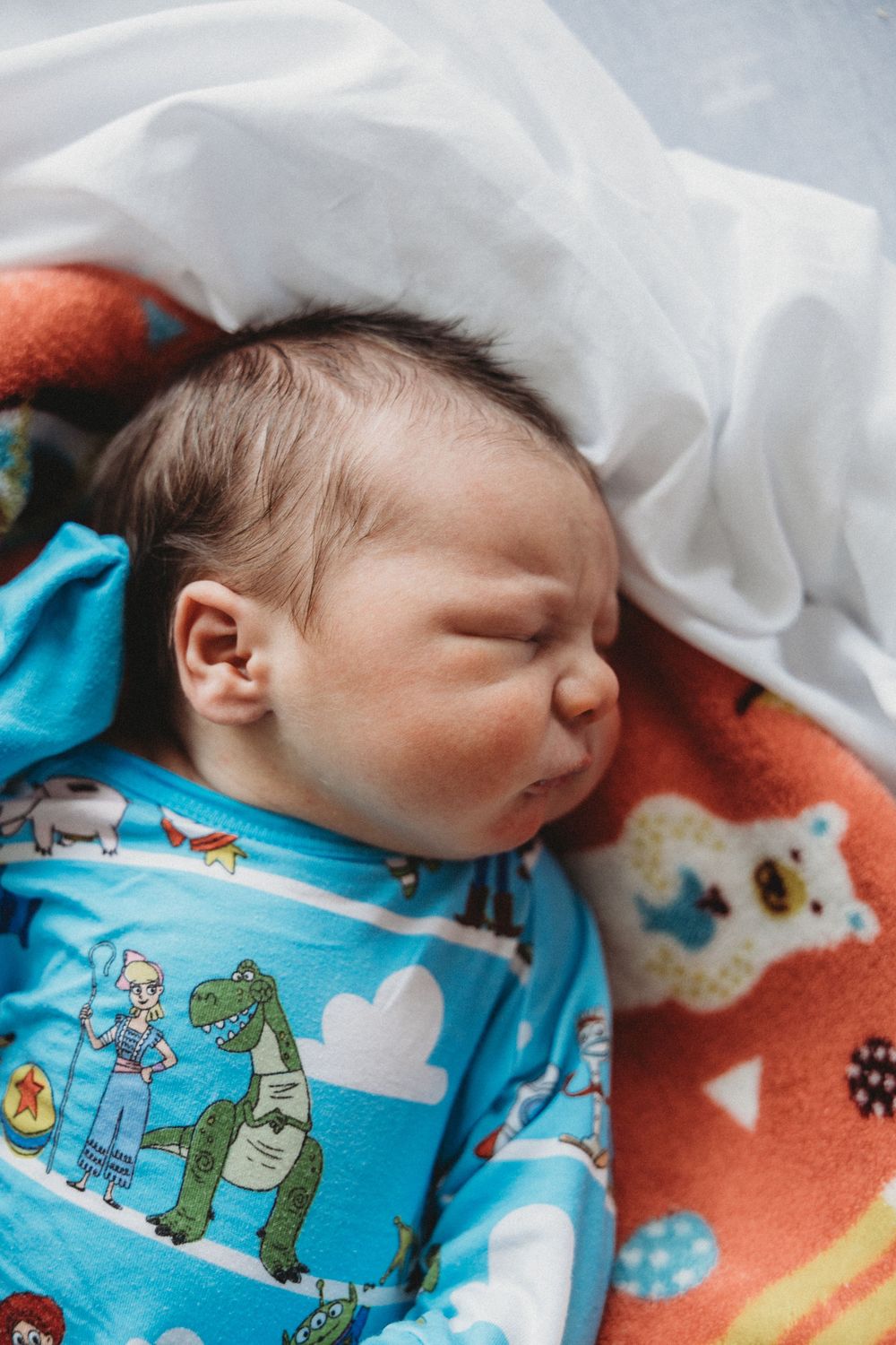 Sleeping newborn baby in blue Toy Story onesie lying on colorful blanket.