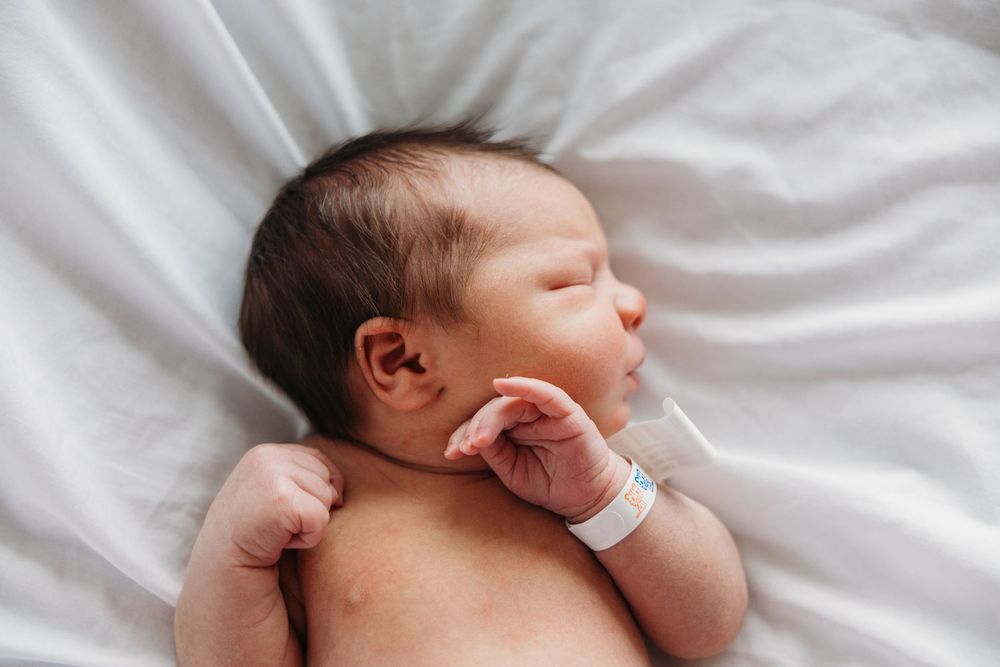 Newborn baby sleeping peacefully on white sheets in hospital with identification bracelet on wrist.