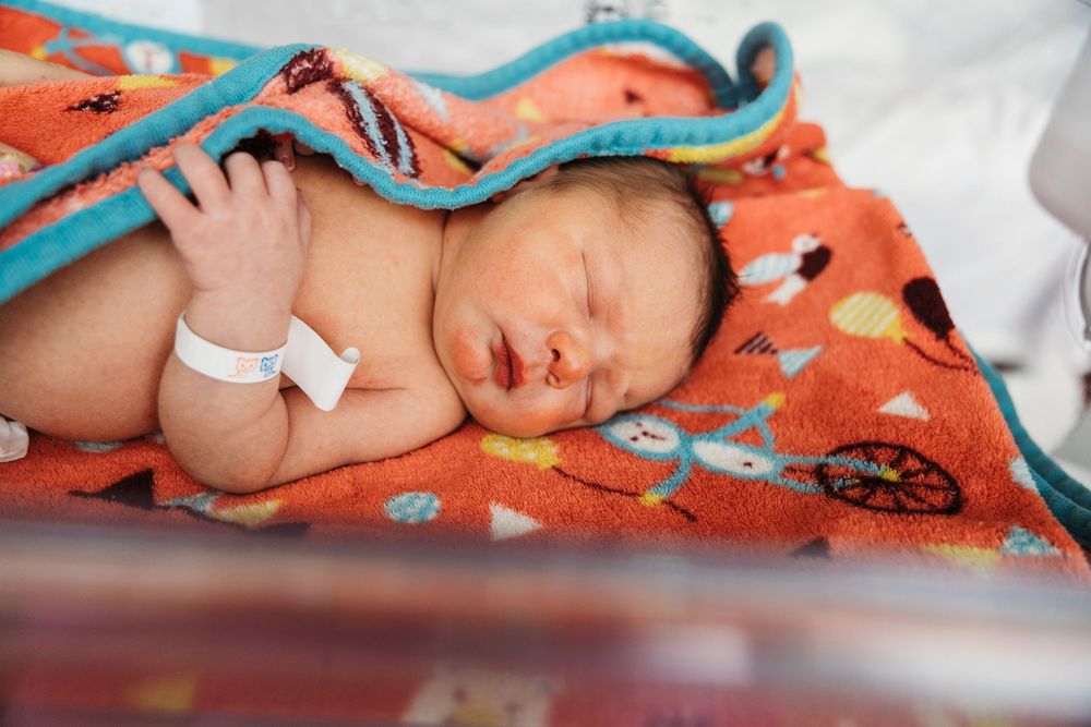 Newborn baby wrapped in colorful orange blanket resting peacefully in hospital bassinet with ID bracelet.