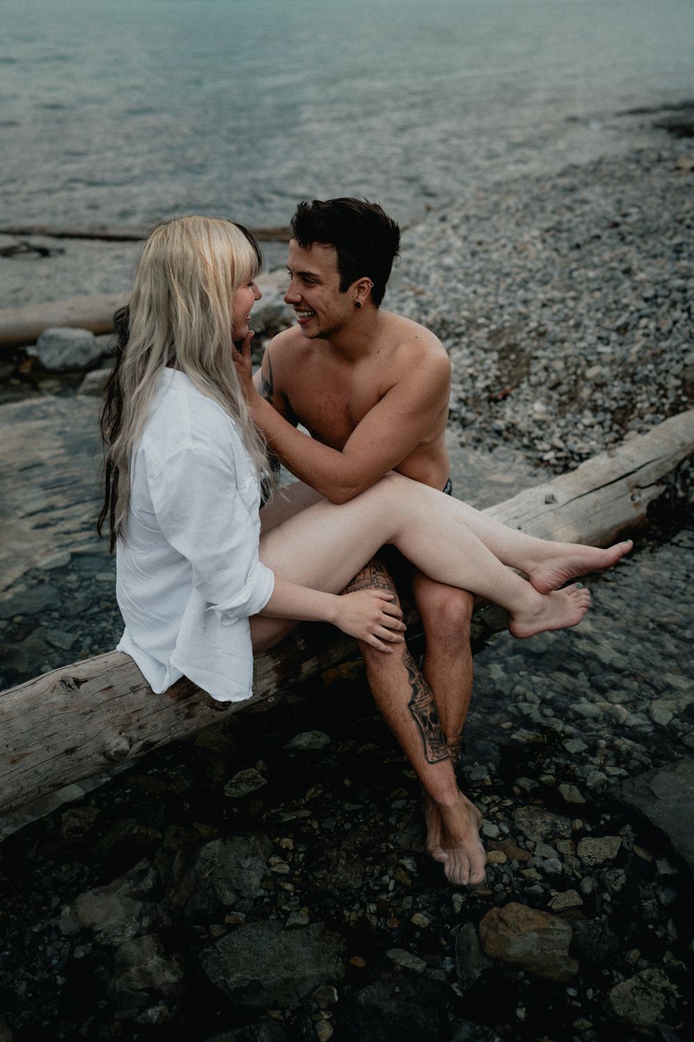 Smiling couple sitting on driftwood log by rocky beach shore near calm water.