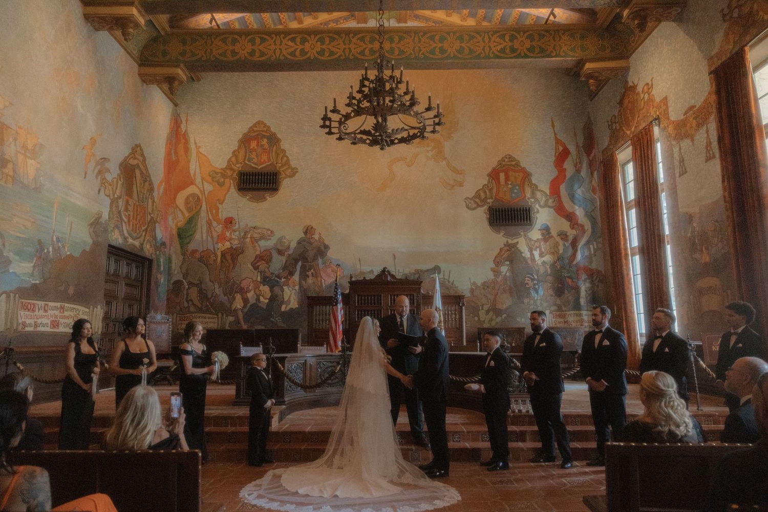 Wedding ceremony in ornate room with murals, bride in white gown with veil, groom, and wedding party at the altar.