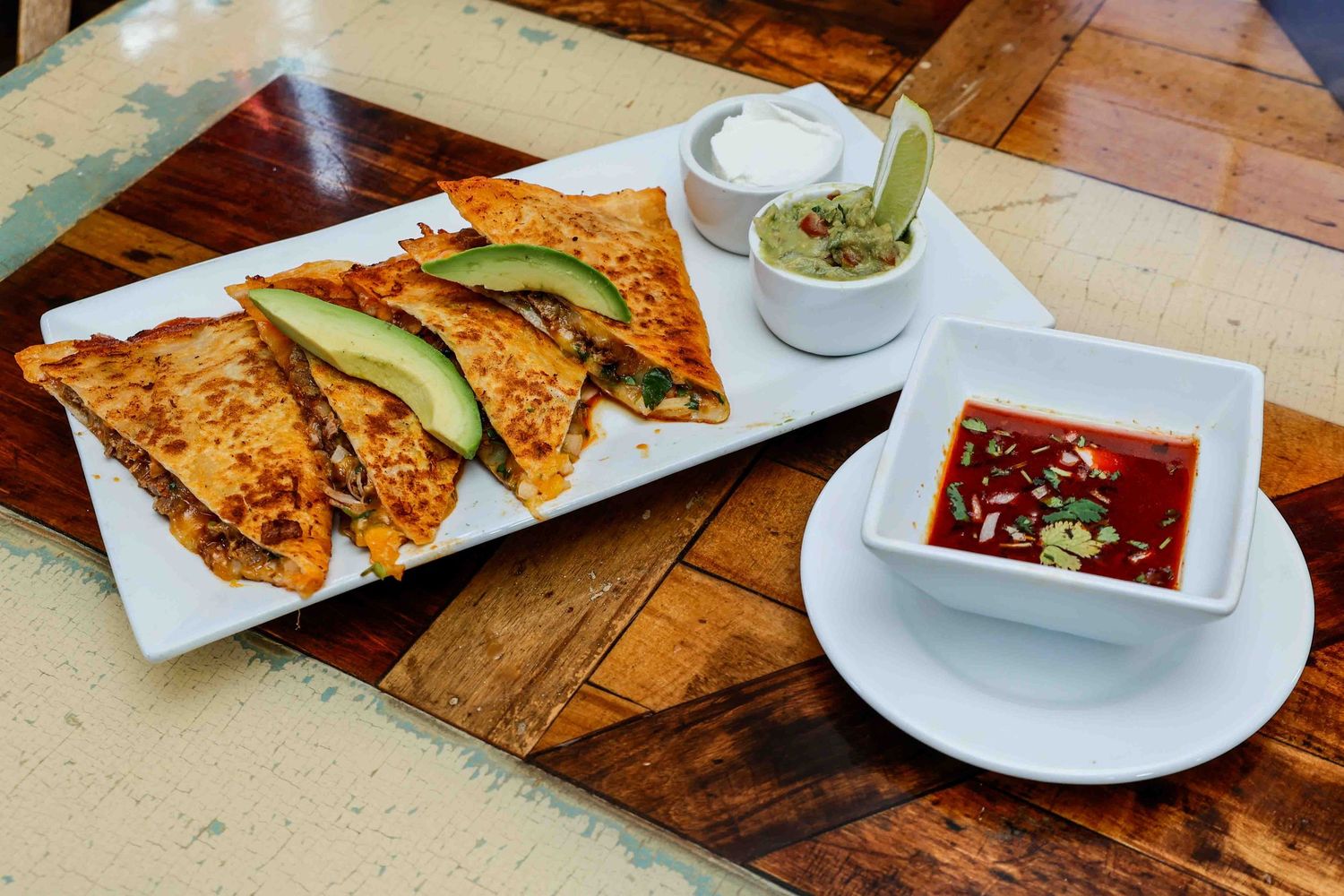Crispy quesadillas with avocado slices on white plate served with salsa dipping sauce and sour cream guacamole sides.