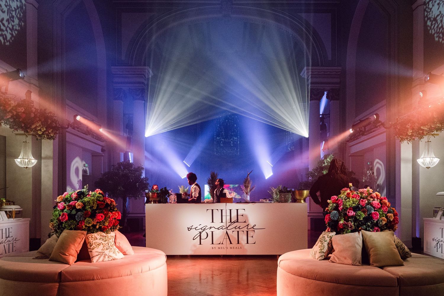 Elegant event bar setup with "The Plate" signage, floral arrangements, dramatic stage lighting in a grand venue.