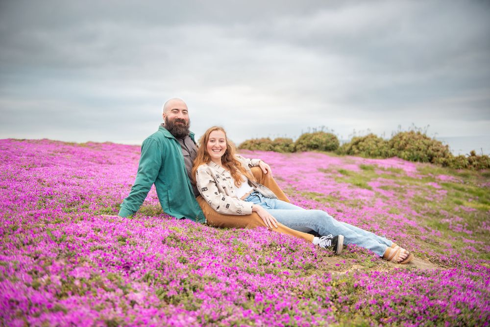 Monterey Engagement photos sitting together in a vibrant pink flower field under cloudy skies, romantic outdoor portrait photography.
