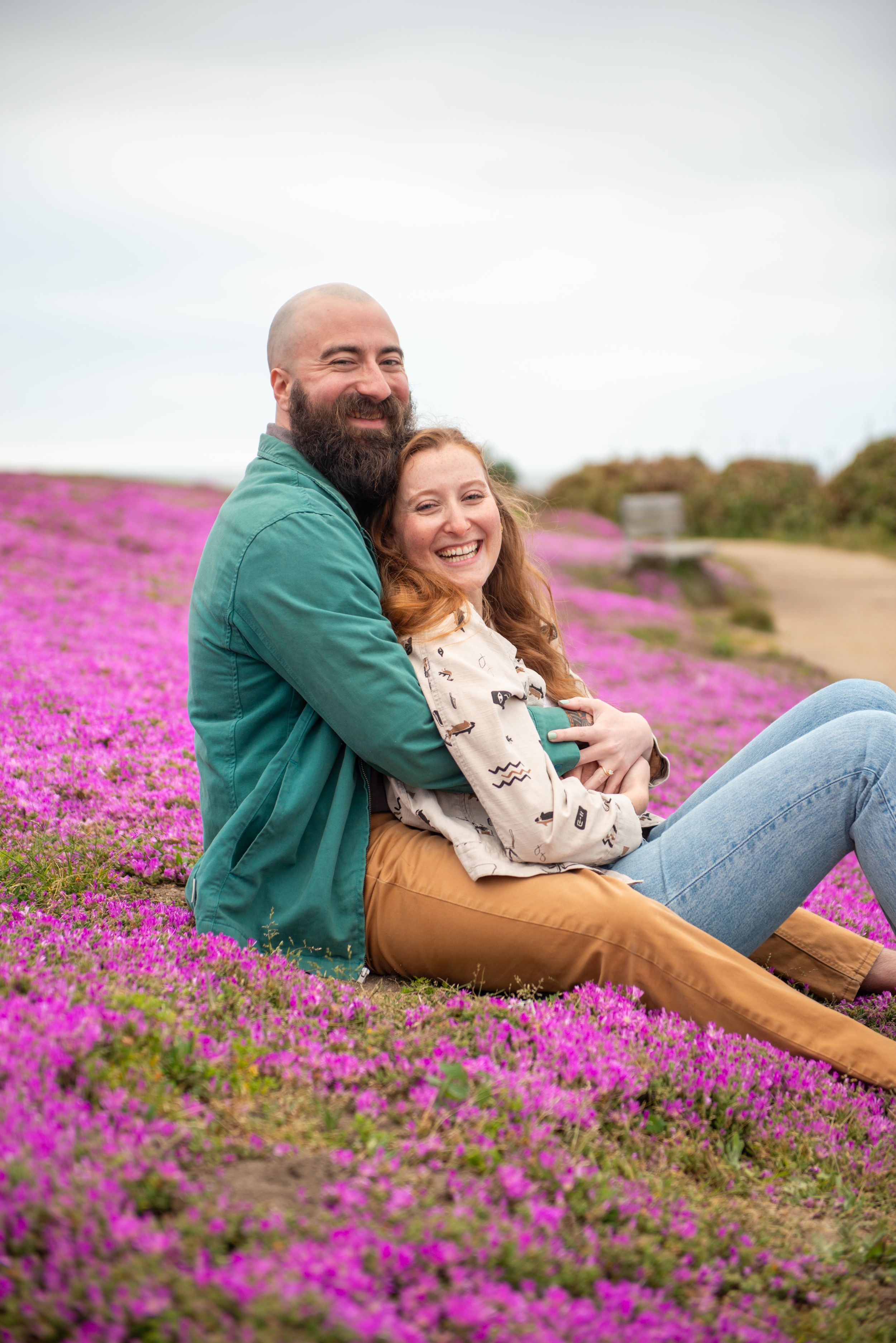 Monterey Engagement photos sitting in a vibrant purple flower field, smiling and embracing, with a scenic path in the background.