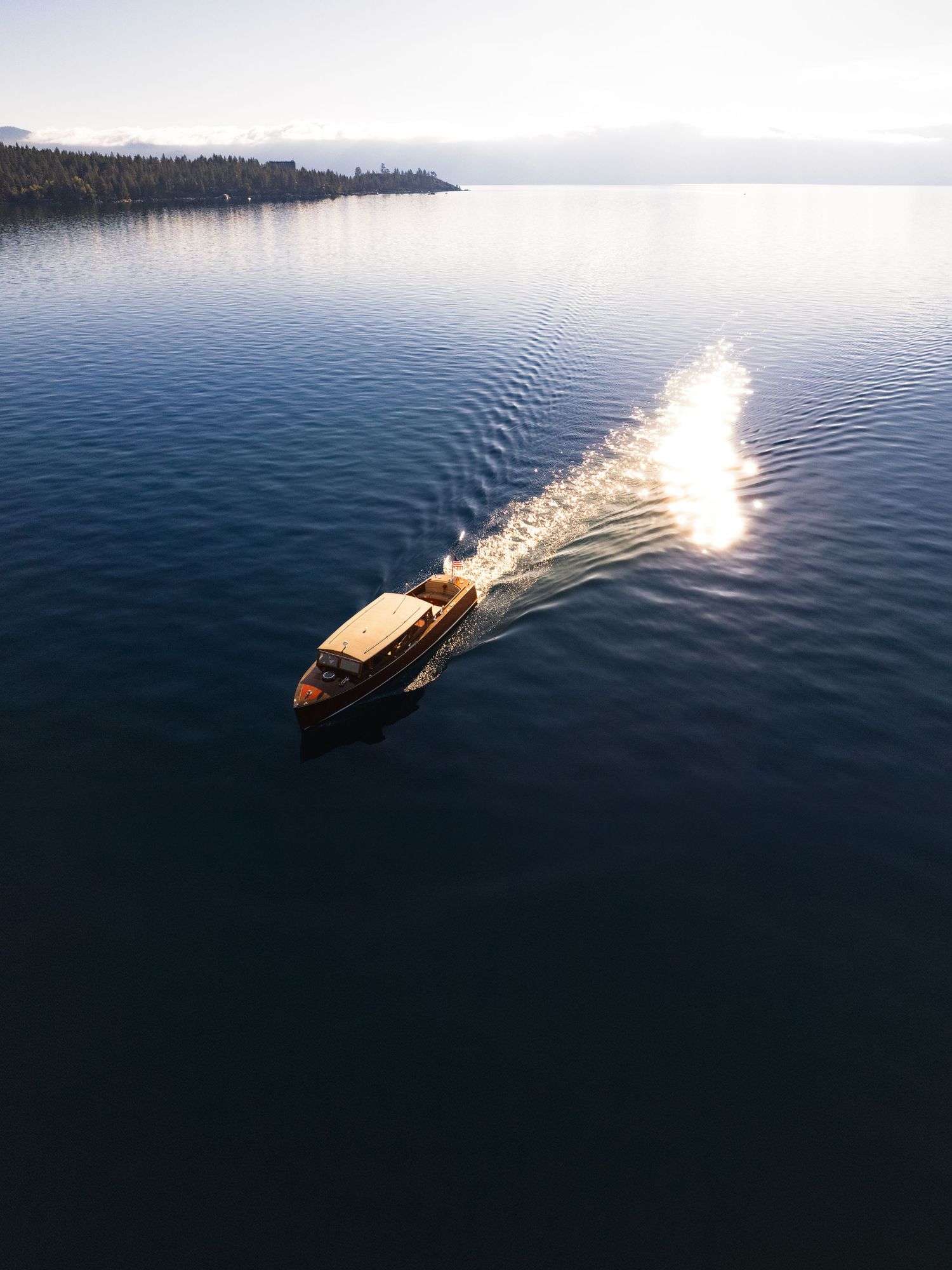 Aerial view of a classic wooden boat cruising on calm blue lake waters, leaving a sparkling wake in golden sunlight.