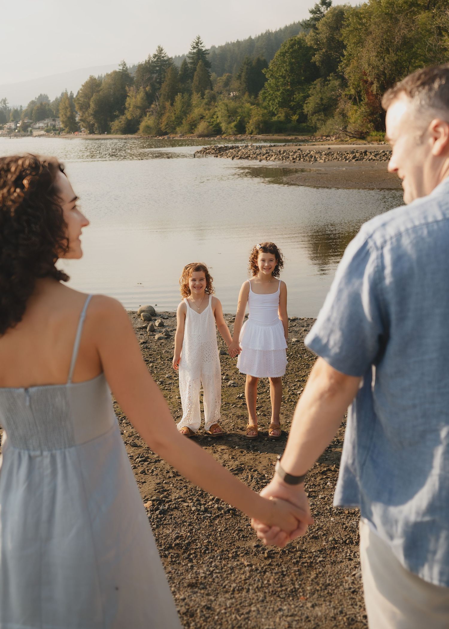 Family holding hands on a rocky beach shore with two young girls in white dresses smiling near a lake.