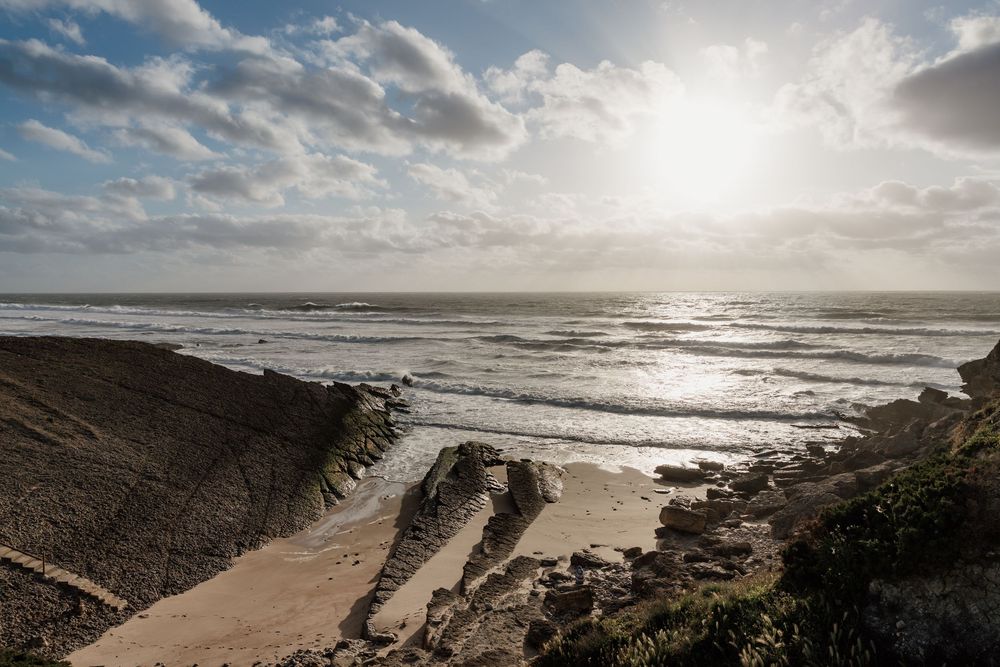 Dramatic coastal landscape with rocky cliffs, sandy beach, and sunlit ocean waves under a cloudy sky.