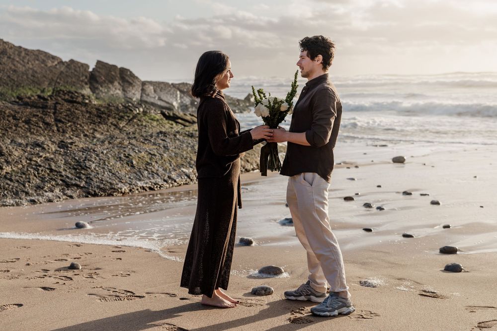 Couple exchanging flowers on a rocky beach at sunset, romantic outdoor proposal scene with ocean waves in the background.