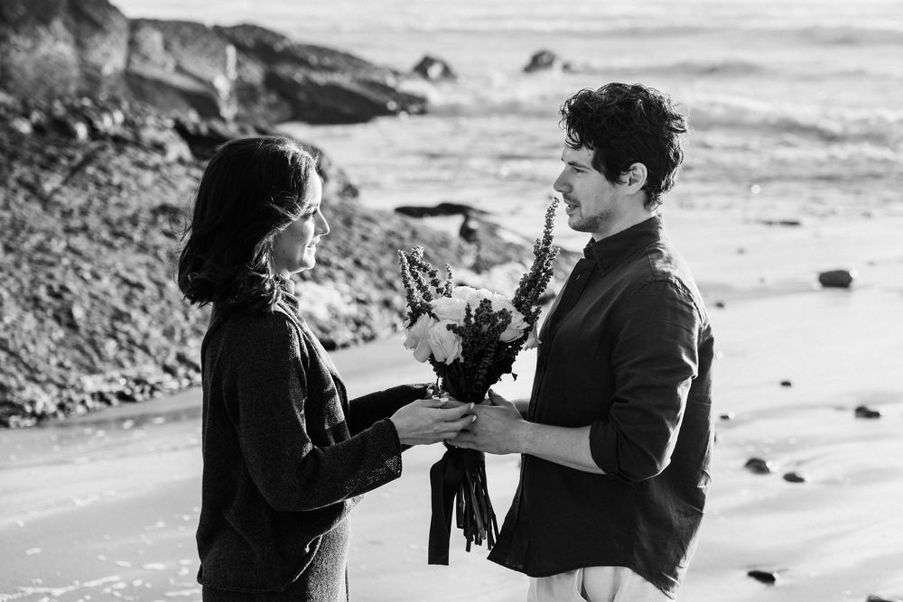 Black and white photo of a couple exchanging flowers on a rocky beach with ocean waves in the background.