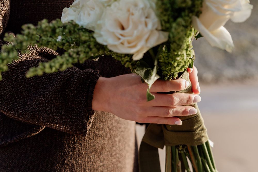 Person holding a bridal bouquet of white roses and greenery wrapped in ribbon, wearing a brown textured jacket.