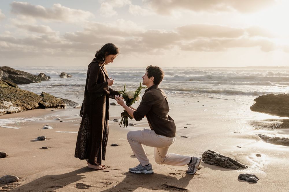 Man proposing to woman on a sandy beach at sunset, kneeling with a rose while ocean waves crash in the background.