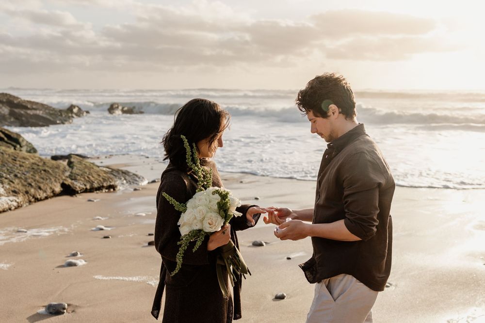 Couple sharing a romantic beach proposal at sunset, man presenting ring to woman holding white bouquet by ocean waves.