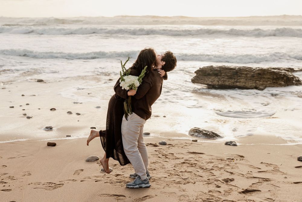 Joyful couple embracing and kissing on a sandy beach with ocean waves and rocks in the background.