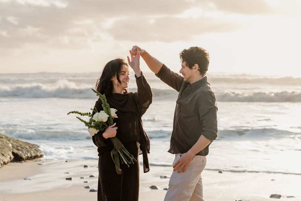 Couple dancing on beach at sunset, woman in black dress holding white flowers while man twirls her near ocean waves.