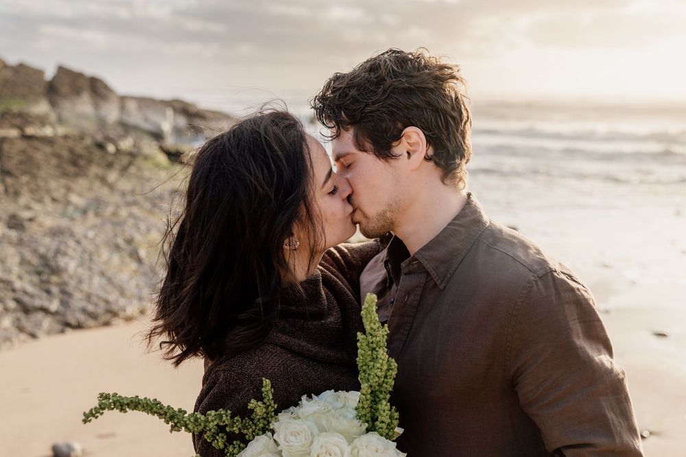Couple sharing a romantic kiss on a beach at sunset, with the woman holding a white floral bouquet.