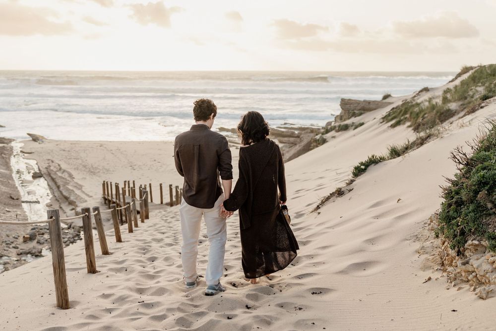 Couple holding hands walking toward the ocean on sandy beach dunes at golden hour sunset.