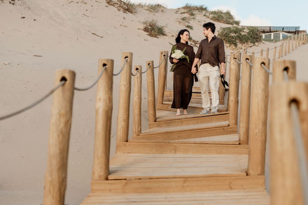 Romantic couple walking hand in hand on a wooden boardwalk through sandy dunes, woman holding a bouquet.