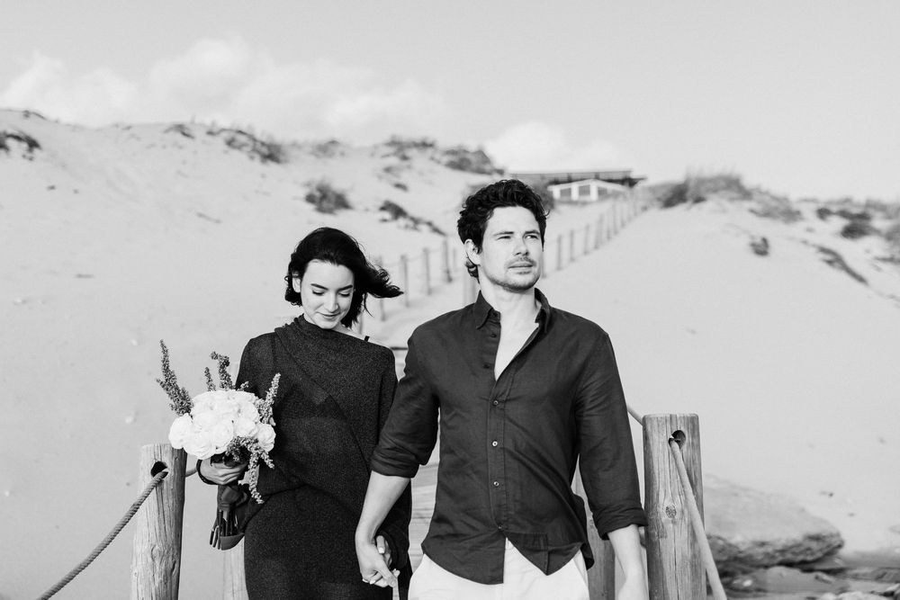 Black and white photo of a couple holding hands walking along a sandy beach path with dunes in the background.