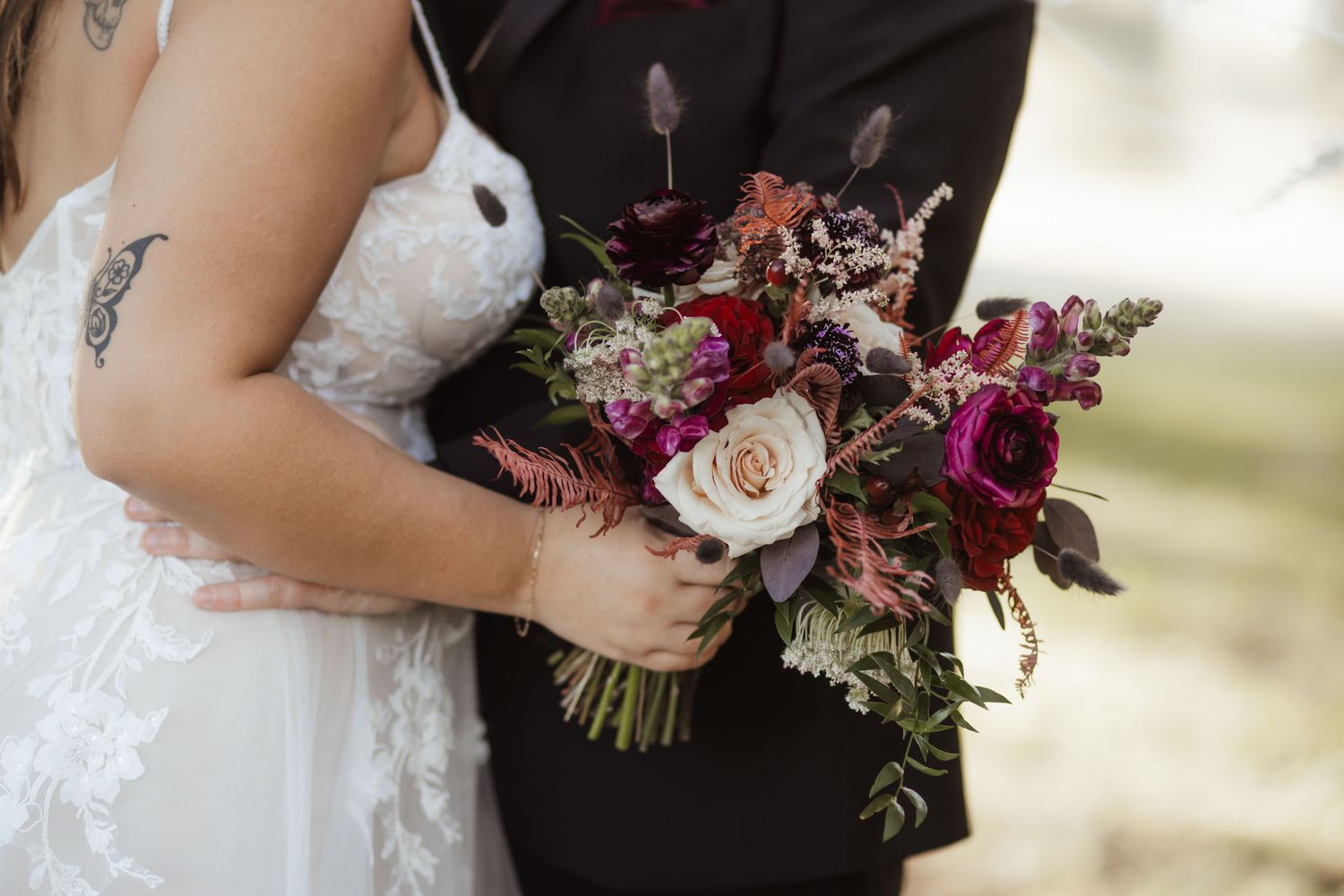 Bride holding a lush burgundy and blush wedding bouquet with dark foliage accents beside groom in black suit.