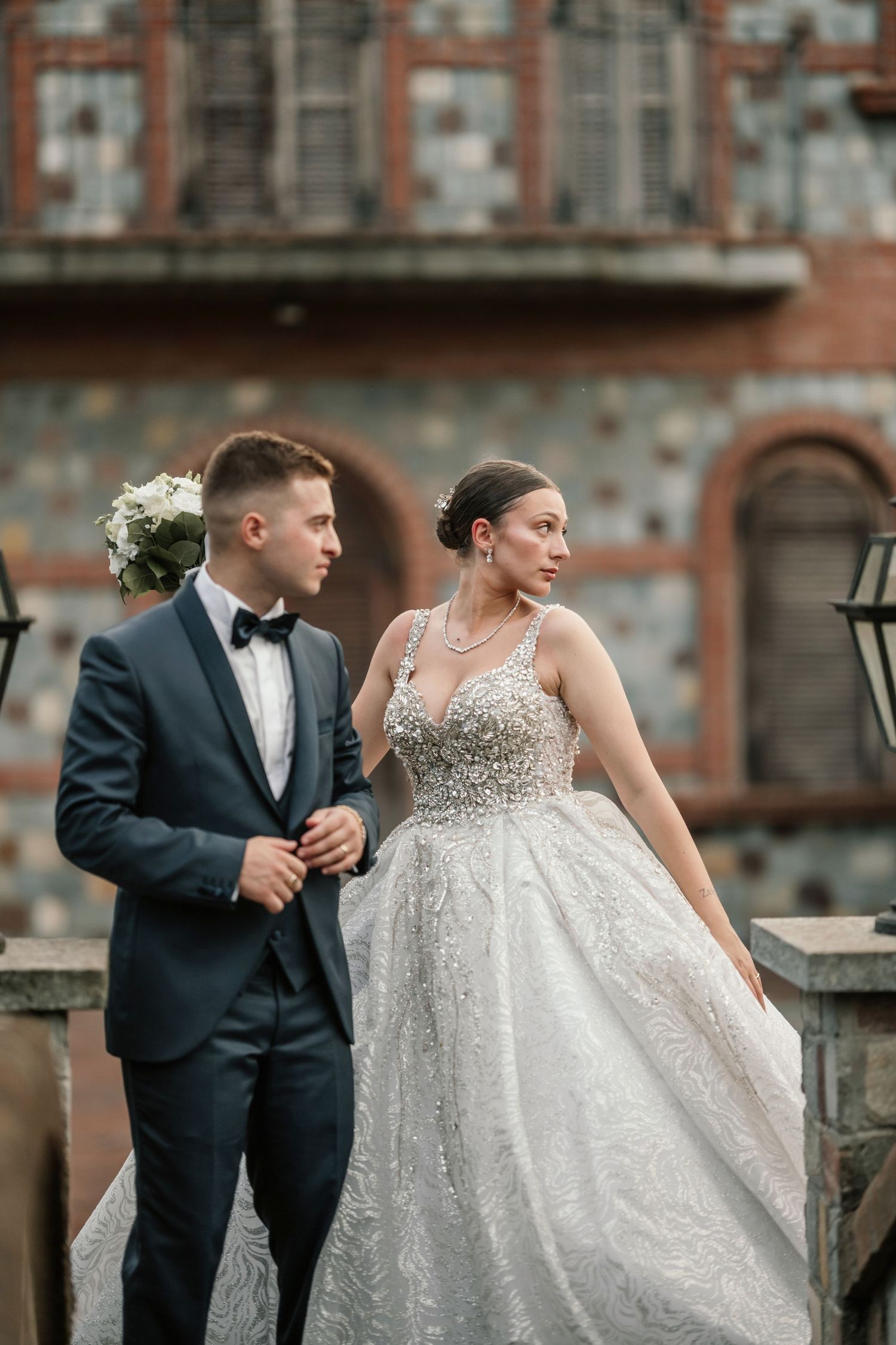 Elegant bride in beaded ball gown and groom in navy tuxedo posing before a stone building.
