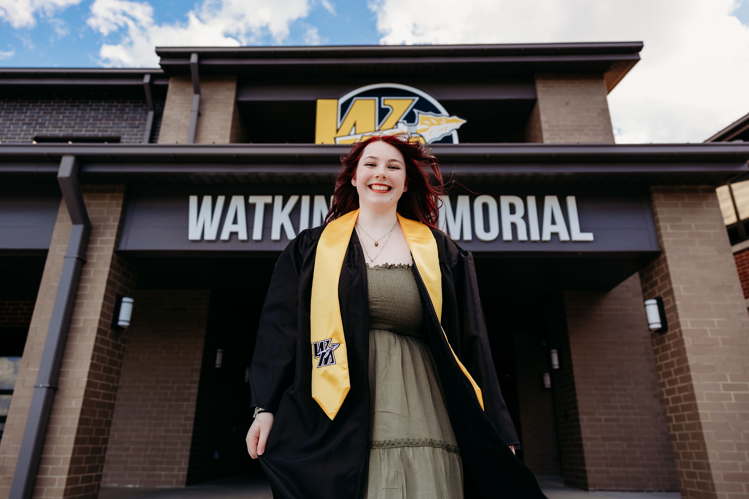 Female graduate in black robe and gold stole smiling in front of Watkins Memorial building.