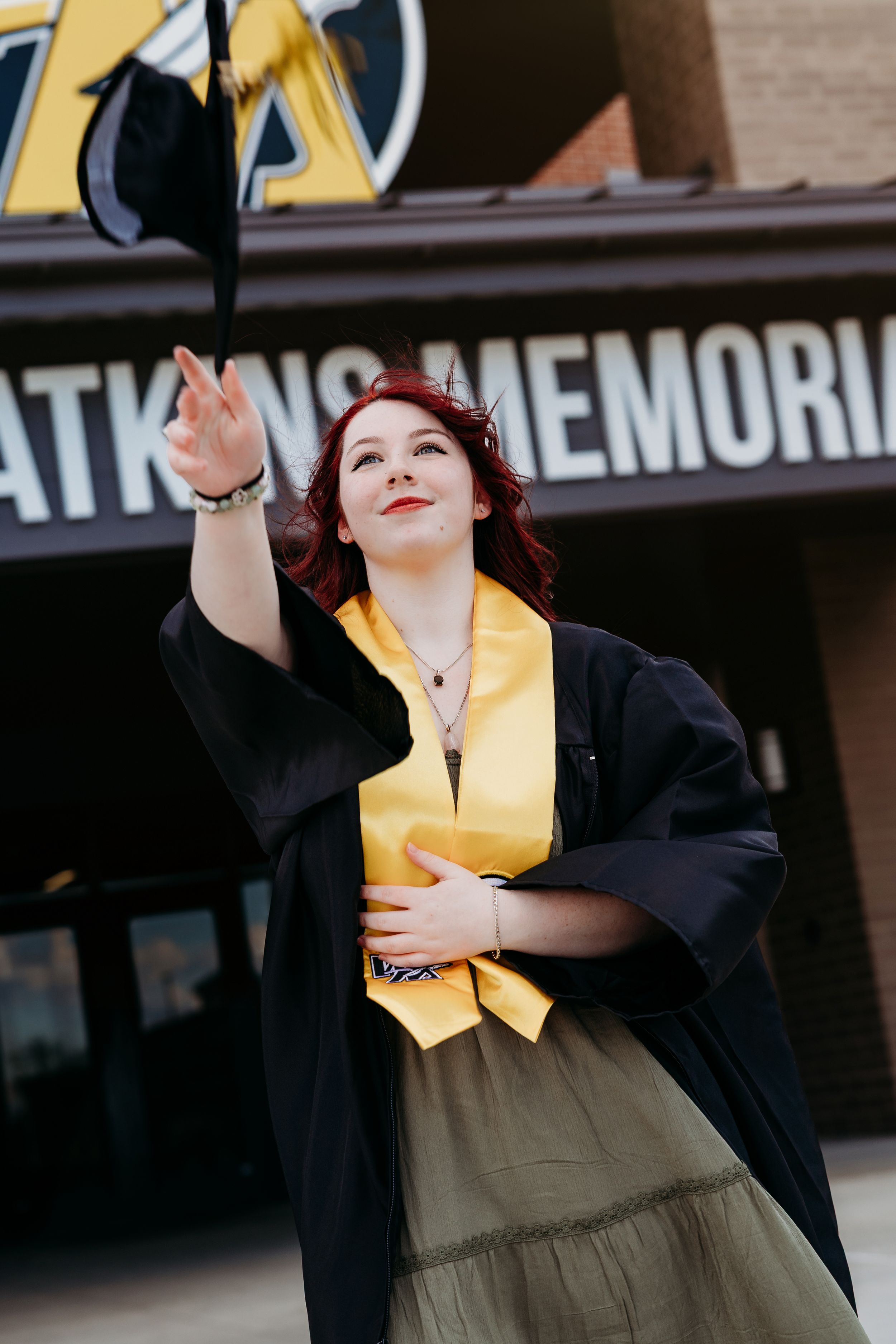 Red-haired graduate in black gown and gold stole tosses cap outside Atkins Memorial building.