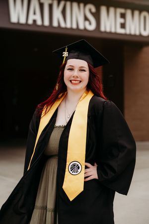Smiling graduate with red hair in black cap and gown with gold stole outside Watkins Memorial.