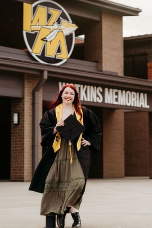 Young woman in black graduation gown and gold stole posing proudly in front of Watkins Memorial building.