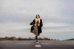 Smiling graduate in black cap and gown with gold stole walking on road under cloudy sky.