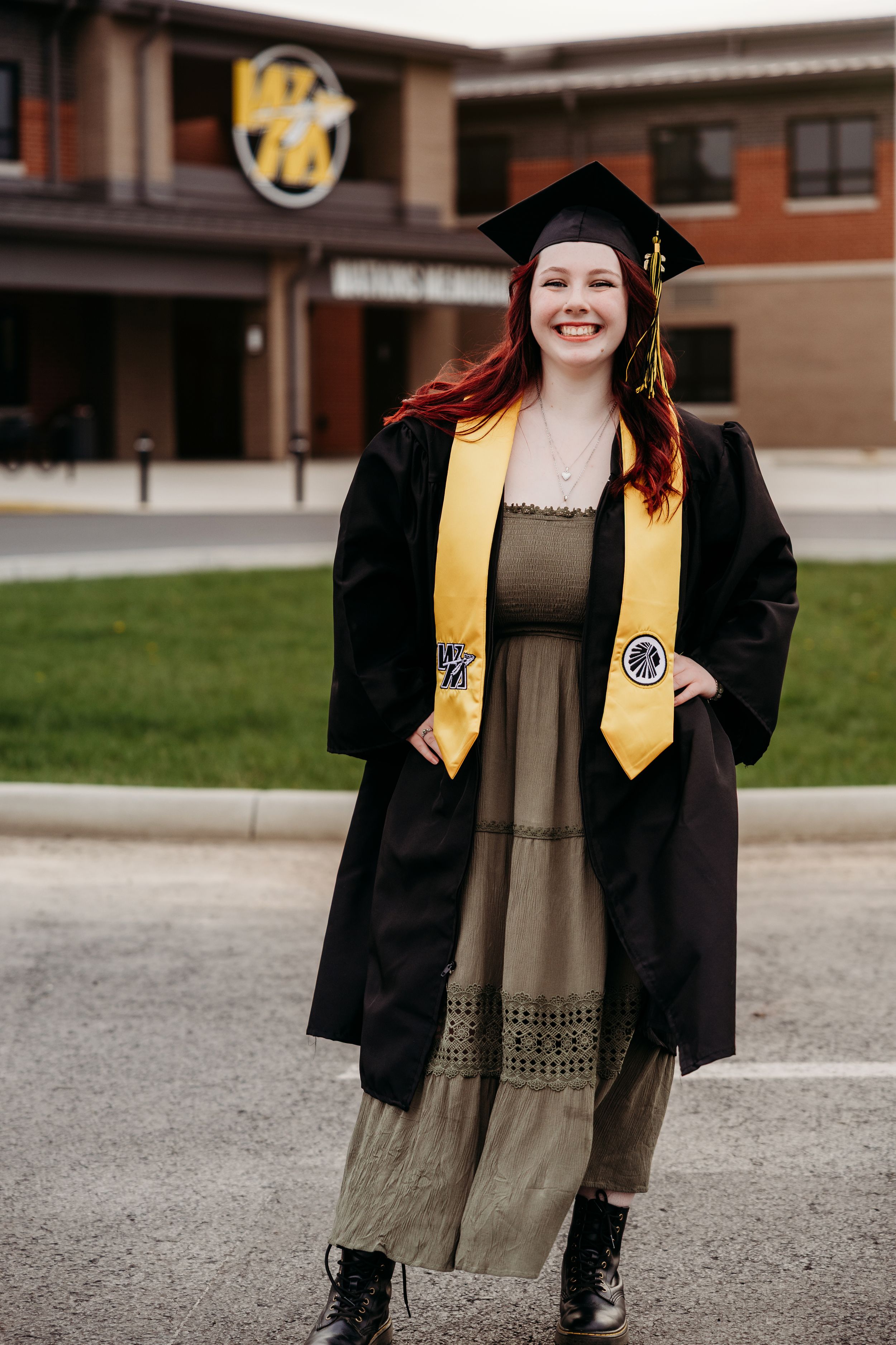 Smiling young woman in black graduation cap and gown with gold stole posing outdoors in front of a school building.